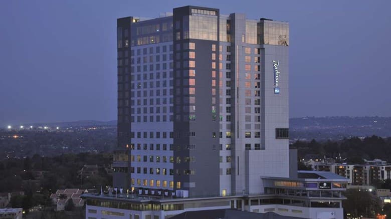A sleek infinity pool on a timber deck at sunset, overlooking the Johannesburg skyline with skyscrapers and green tree canopy in the distance.