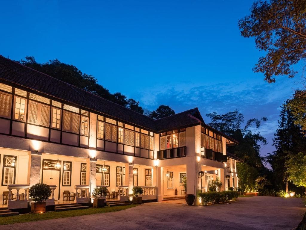 A black and white colonial villa with terracotta roof tiles, viewed from a low angle through lush tropical palm fronds and jungle greenery.