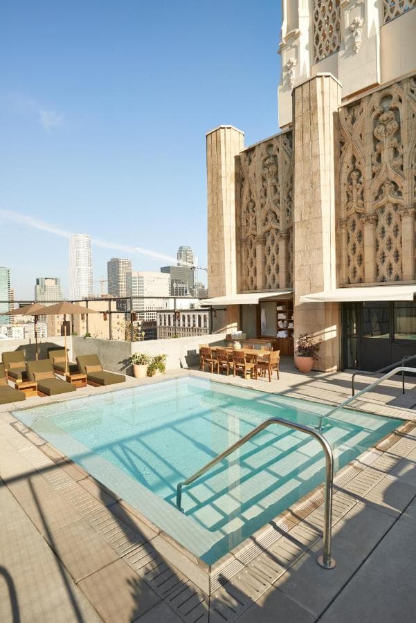 The iconic rooftop concrete plunge pool of STILE Downtown LA at twilight, illuminated by the red 'JESUS SAVES' neon sign against the city skyline.