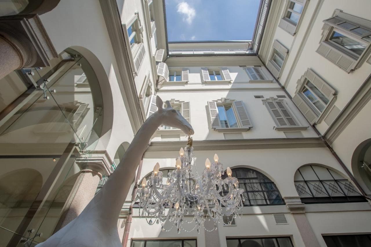 The glass-enclosed courtyard of Hotel Indigo Milan featuring the life-sized 'Giraffe in Love' chandelier by Qeeboo holding a classic chandelier in its mouth.