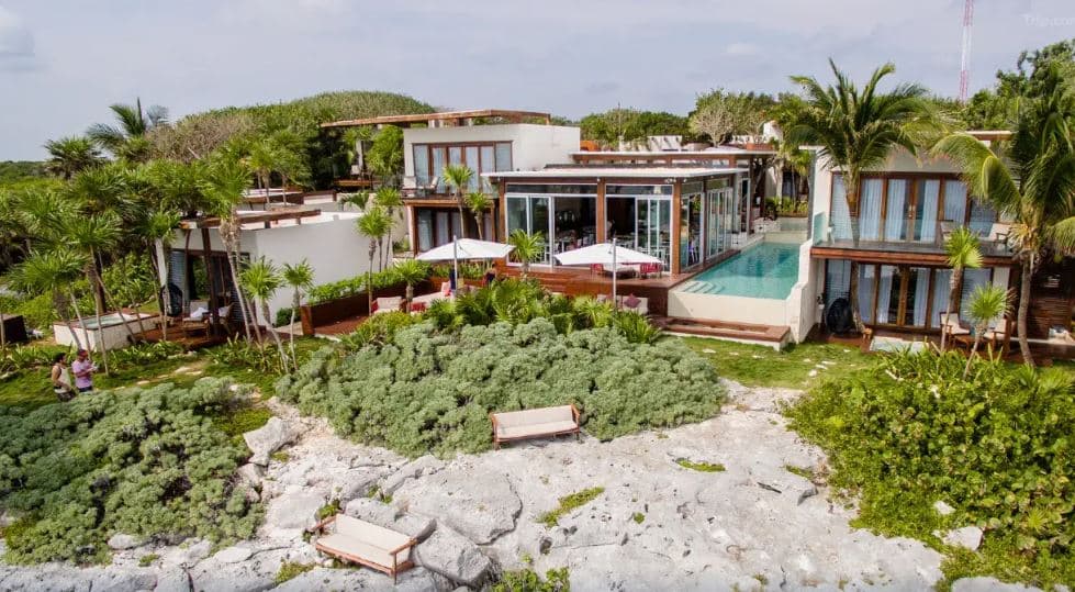 A high-angle shot of the angular infinity pool at Mi Amor Tulum, featuring pastel pink floating daybeds and the crashing Caribbean waves against the rocky cliffside.