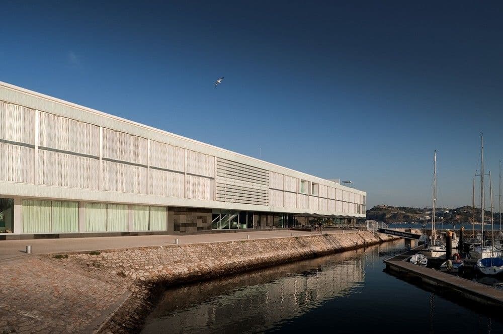 The sleek, white exterior of Altis Belém Hotel & Spa reflecting in the Tagus River at sunset, with the 25th of April Bridge in the background.