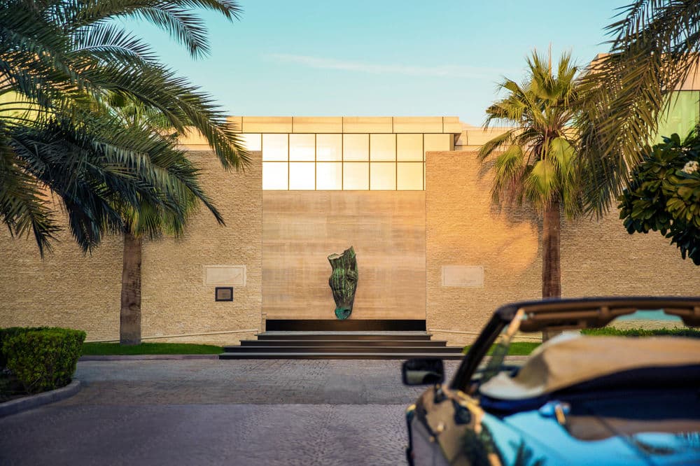 A symmetrical view of the Meliá Desert Palm infinity pool leading the eye toward green polo fields, flanked by palm trees under a clear Dubai sky.