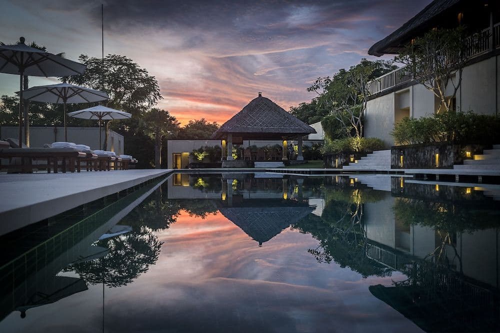 A symmetrical view of the REVĪVŌ main pool framed by stone colonnades and white bougainvillea, reflecting the serene, adult-only atmosphere.