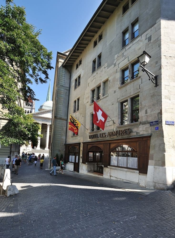 The historic stone facade of Hotel Les Armures in Geneva's Old Town, adorned with Swiss flags and red geraniums.