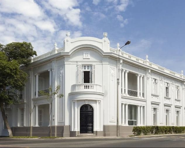 A white Belle Époque mansion facade with columns and open balconies on a corner street in Lima, featuring a cyan sky.