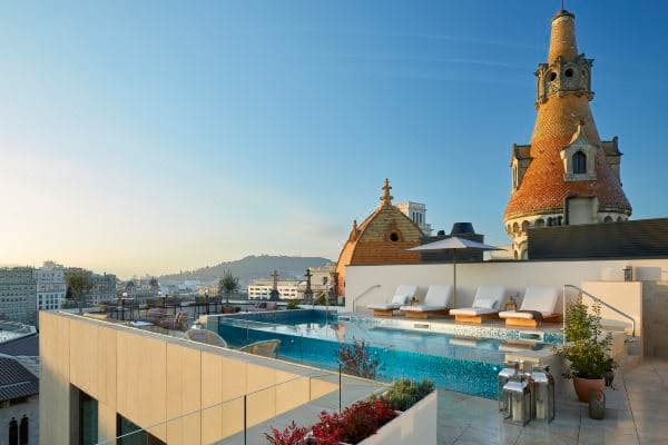 A chic, dimly lit rooftop bar at ME Barcelona at dusk, with guests holding cocktails and the illuminated spires of the Sagrada Família visible in the distance.