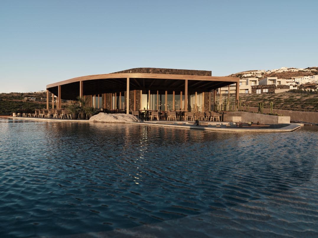 검은 화산석 건축물과 먼 포도밭으로 둘러싸인 Magma Resort Santorini의 거대한 인피니티 풀이 하늘을 반사하는 모습.