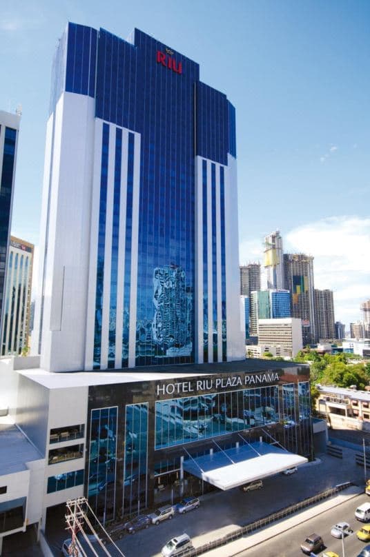 A low-angle shot of the Hotel Riu Plaza Panama's outdoor pool deck, surrounded by gleaming modern skyscrapers under a tropical sky.