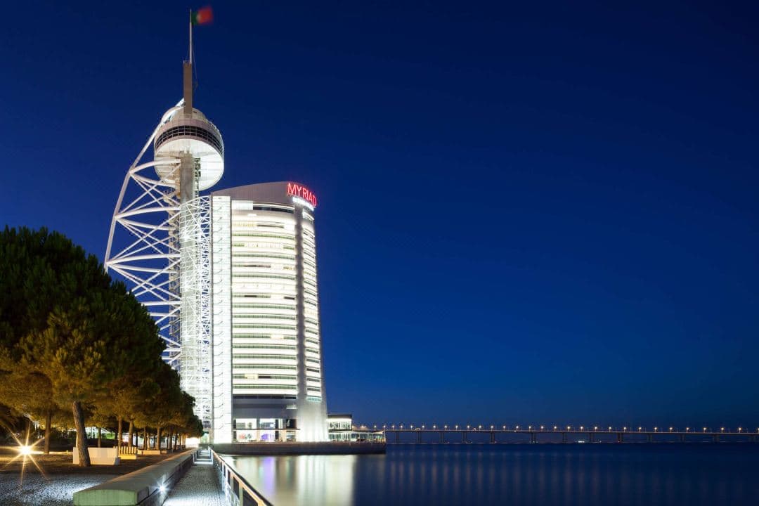 The sail-shaped glass tower of Myriad by SANA rising directly from the Tagus River in Lisbon, illuminated against a twilight sky.
