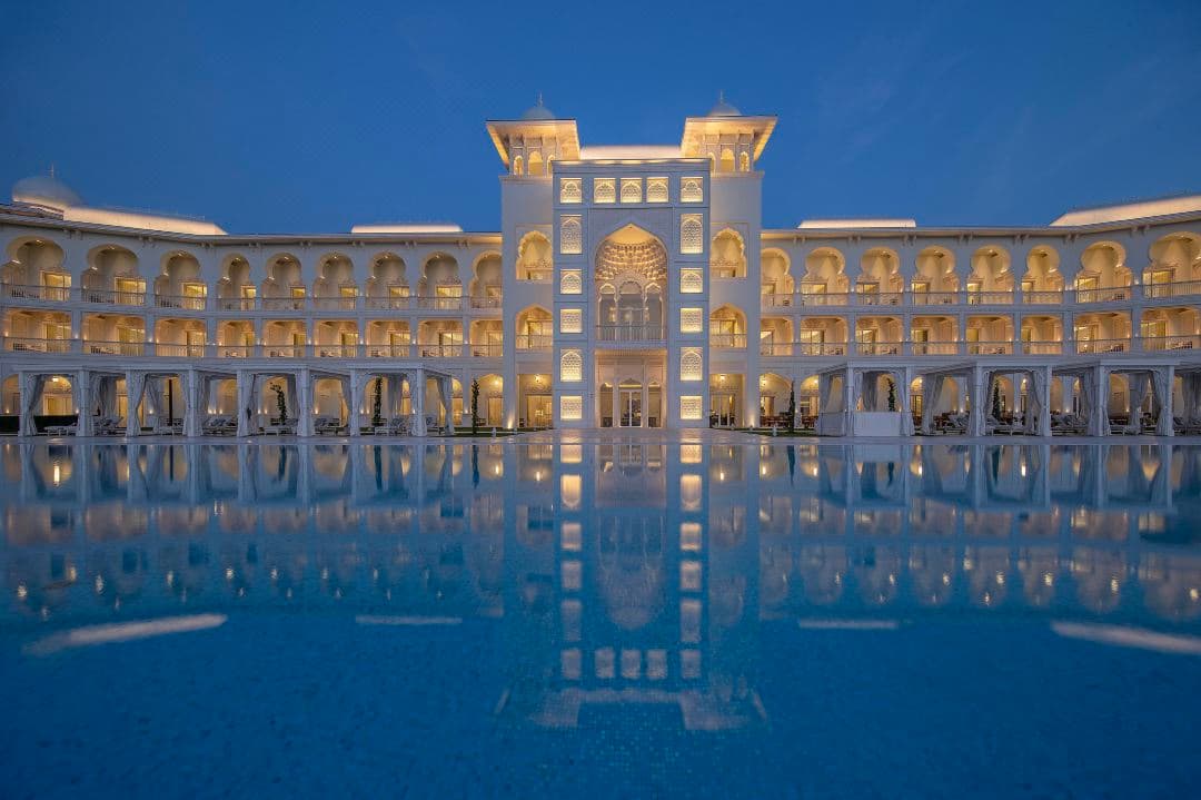 The 60-meter infinity pool at The Chedi Katara, flanked by white Mughal-style arches and palm trees under a blue sky.