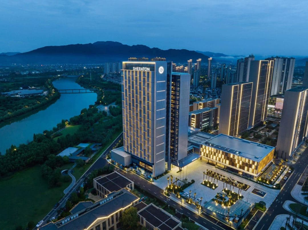 View of the Sheraton Taizhou guest room featuring floor-to-ceiling windows overlooking the Yongning River and Wudong Bridge at dusk.