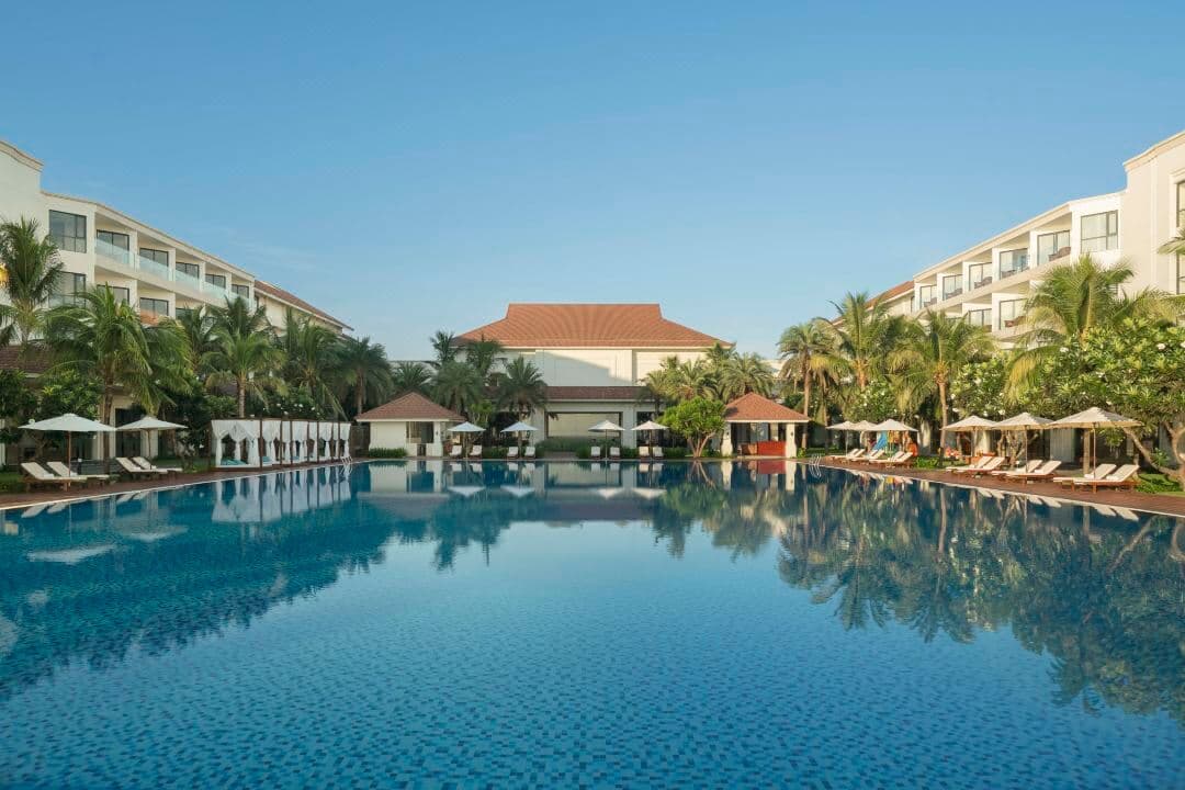 A massive infinity pool at Renaissance Hoi An seamlessly blending with the Thu Bon river estuary and the sea beyond, framed by modern lounge chairs.