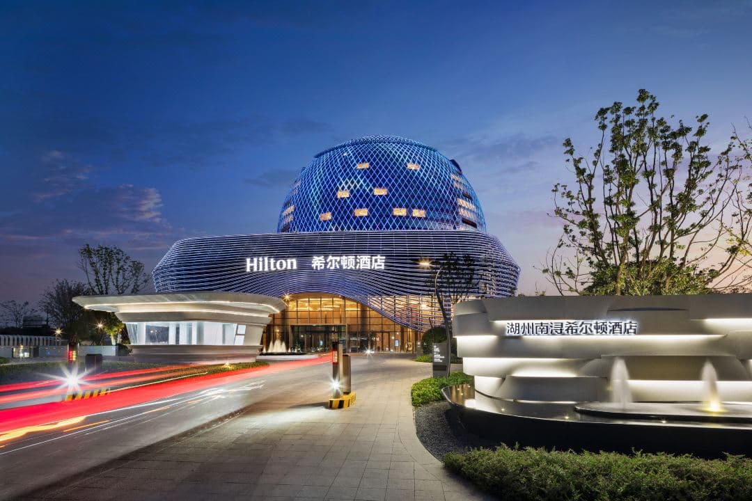 The golden spherical exterior of Hilton Huzhou Nanxun reflecting on Shen Jiayang Lake at dusk.