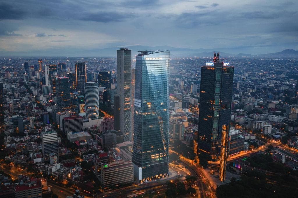 A dramatic, low-angle shot of The Ritz-Carlton Mexico City's glass facade reflecting the sunset, with the iconic Chapultepec Park tree canopy visible below.
