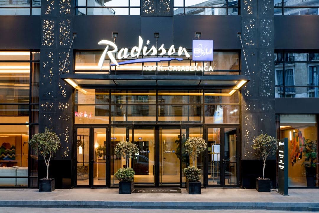 Sleek glass facade of Radisson Blu Casablanca contrasting with historic Art Deco buildings, sunset reflection on windows.