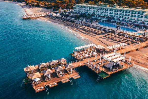 Triangular bamboo cabanas on a wooden pier at Corendon Playa Kemer, framed by turquoise water and pine-covered mountains.