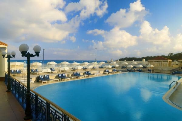 A view from a high hotel balcony looking out over the lush Montazah Palace gardens and the Mediterranean Sea at sunset.