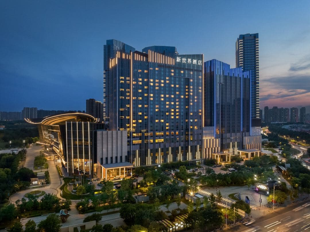 A sleek, modern hotel room at New World Shenyang with floor-to-ceiling windows overlooking the Hun River and Shenyang Grand Theatre at dusk.