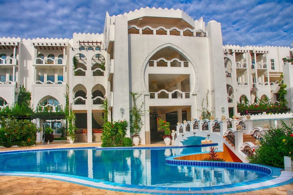 The white arched facade of Madinat Al Bahr framed by palm trees, reflecting into the expansive outdoor swimming pool at twilight.