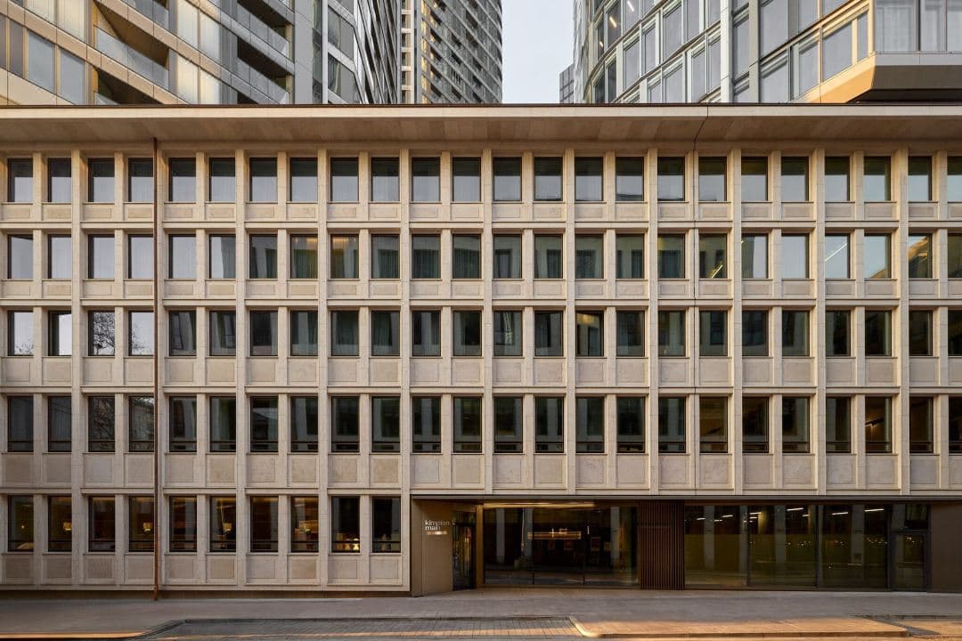 A low-angle shot of the Kimpton Main Frankfurt's facade, featuring a 1950s sandstone exterior contrasting with modern turquoise window accents.