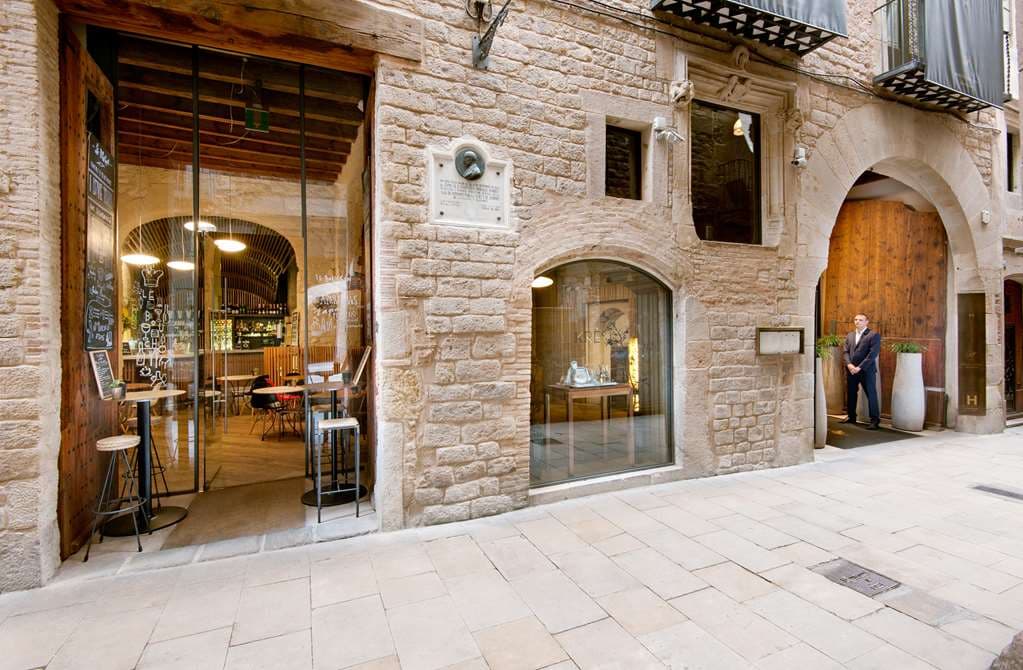 Sunlight filtering through the ancient arches of the Mercer Hotel Barcelona's Patio de los Naranjos, illuminating the orange trees and Roman stone walls.