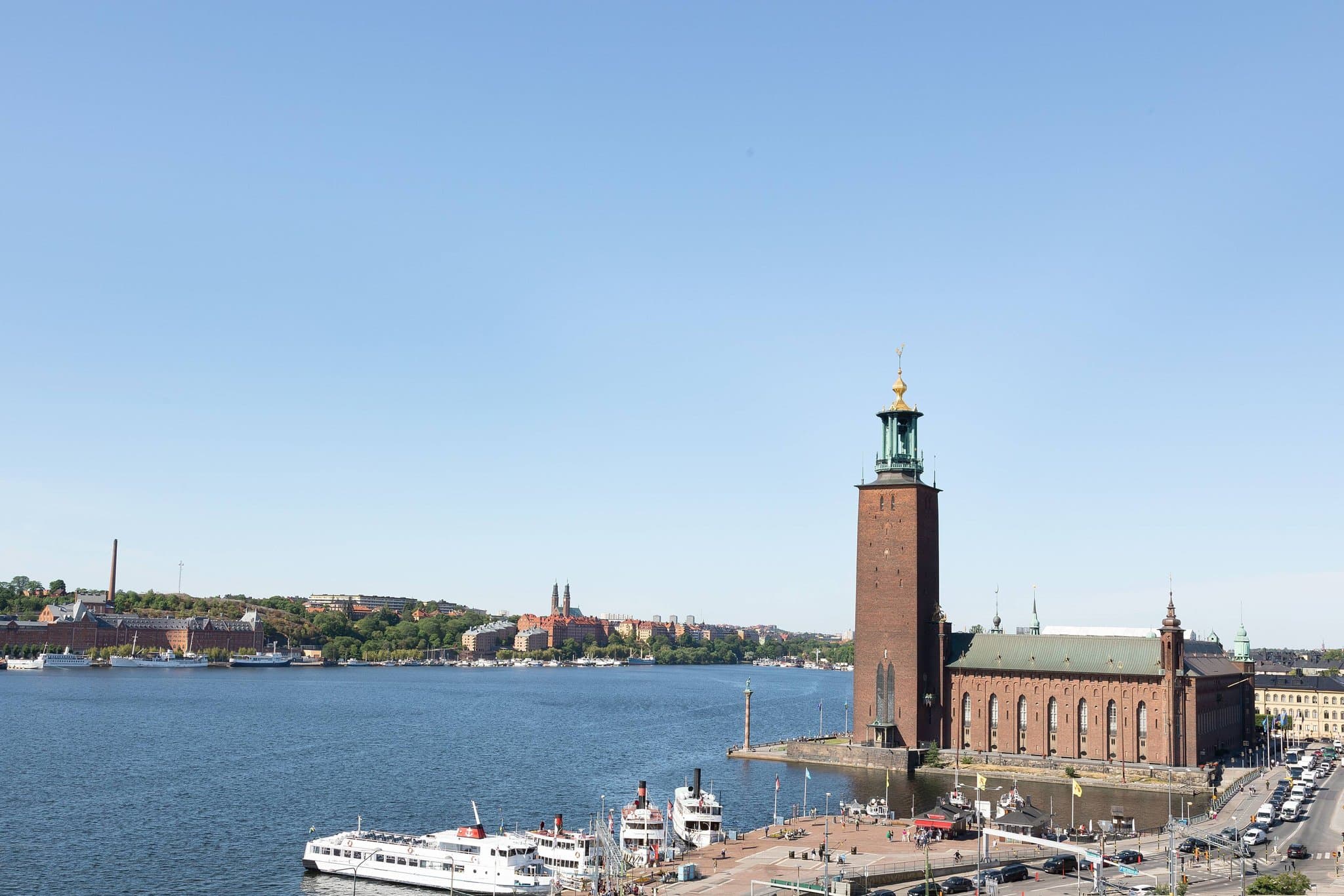 A spacious hotel room with a private balcony overlooking Stockholm City Hall and Lake Mälaren at twilight.