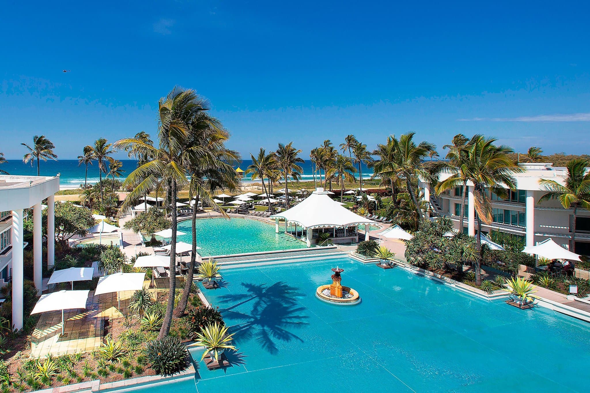 View of the Sheraton Grand Mirage lagoon pool winding towards the ocean, framed by palm trees and white resort architecture.