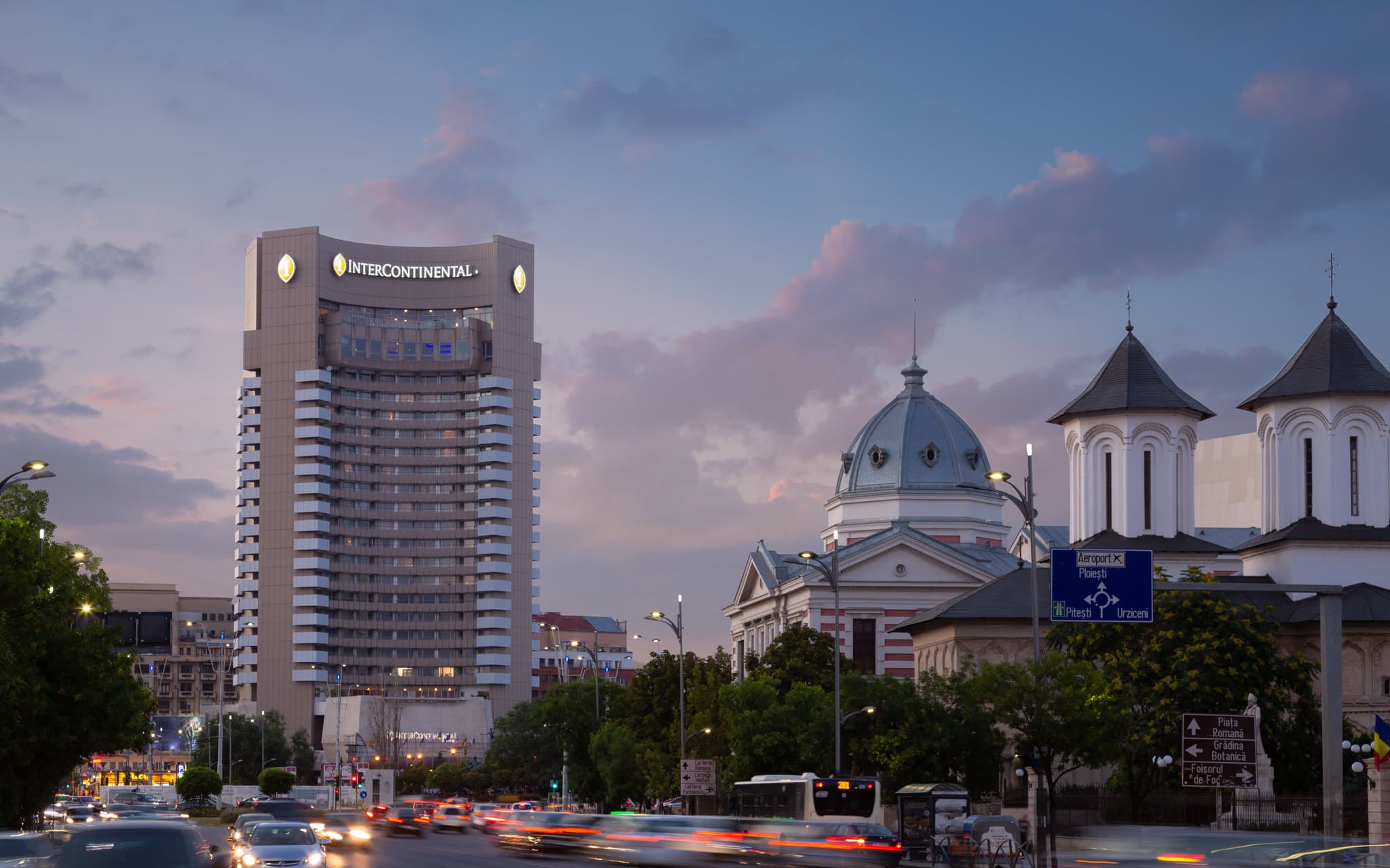 A dramatic low-angle shot of the Grand Hotel Bucharest's towering concrete facade at dusk, with lights glowing from the unique curved balconies.