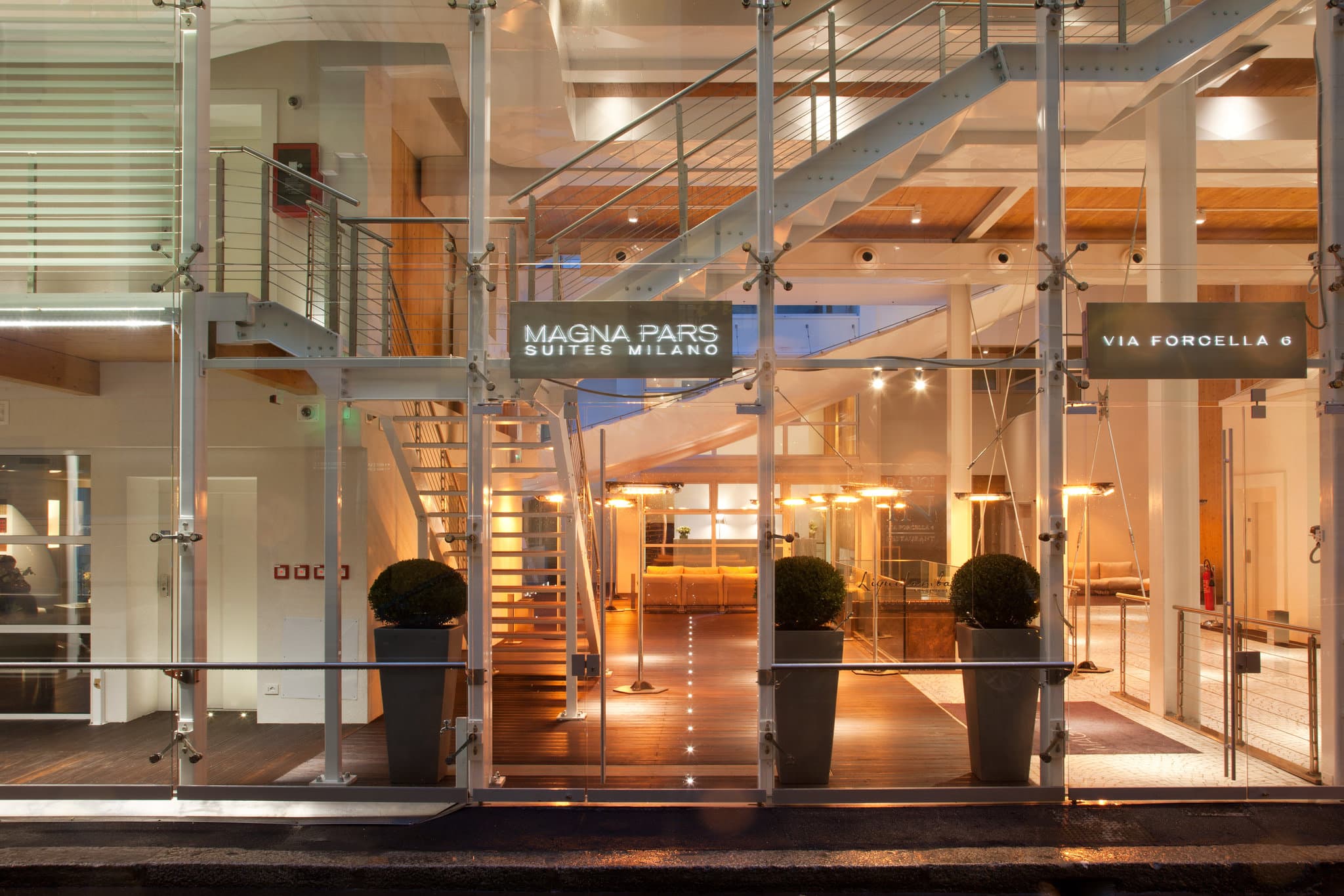 The lush internal courtyard of Magna Pars l'Hotel à Parfum viewed through floor-to-ceiling glass windows, featuring white modern furniture and abundant greenery.