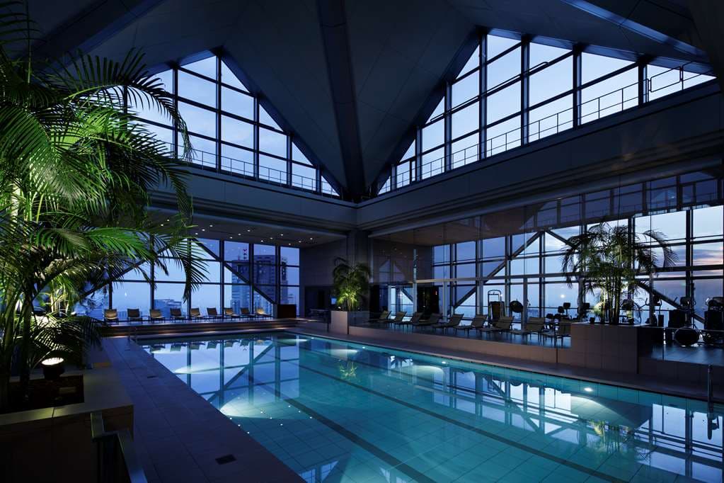 A dramatic view of the Park Hyatt Tokyo's 47th-floor swimming pool under a glass pyramid atrium at twilight, with the Tokyo city lights visible below.