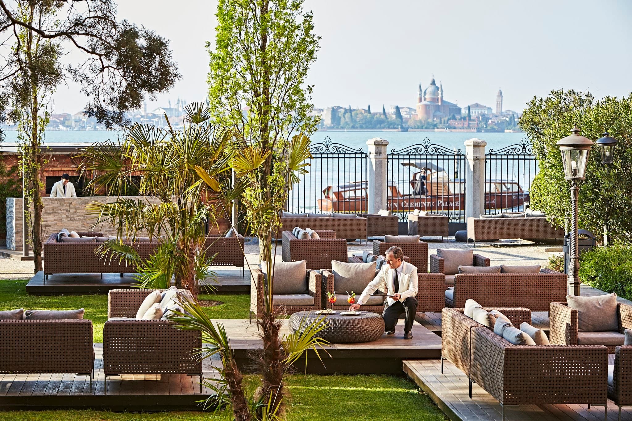 A manicured private island garden in Venice featuring a large heated swimming pool surrounded by white cabanas, with the historic San Clemente church facade in the background.