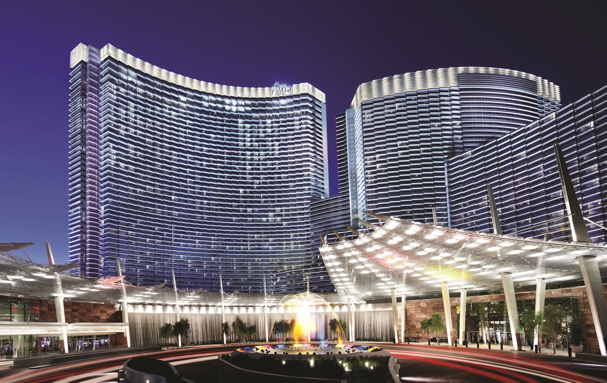 A panoramic view of the Las Vegas Strip at night seen through the floor-to-ceiling corner windows of an ARIA Sky Suite, featuring a chaise lounge and modern decor.