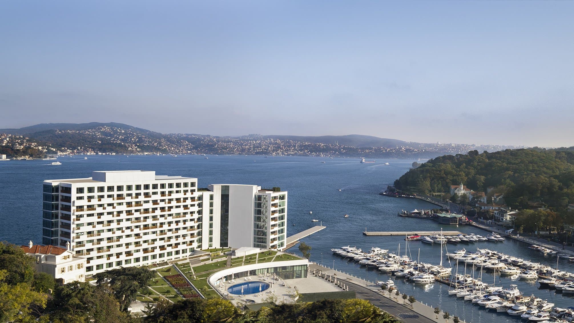 A guest sipping espresso on a private glass-fronted balcony at The Grand Tarabya, overlooking a passing ship on the turquoise Bosphorus strait.