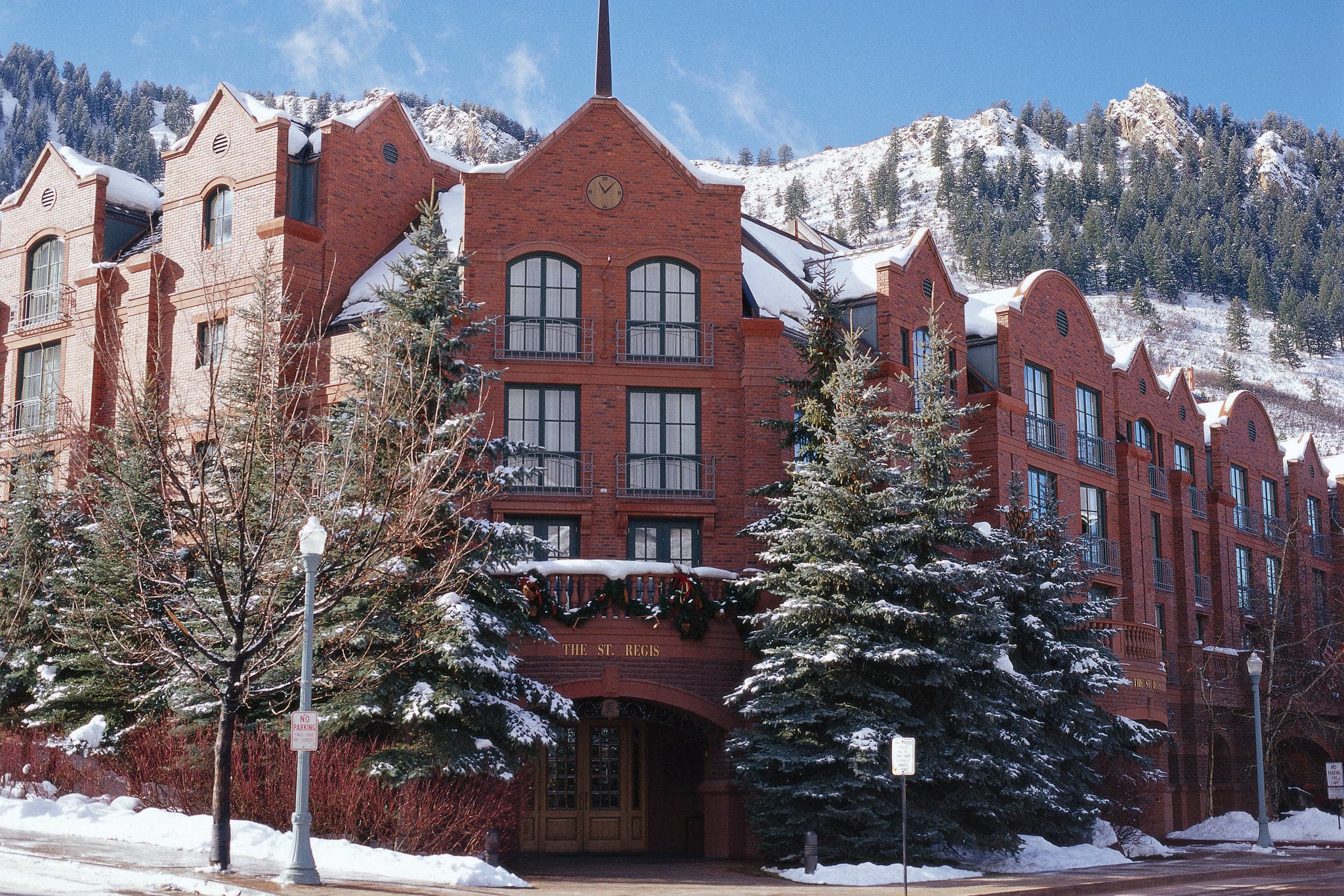 A snowy outdoor courtyard at St. Regis Aspen featuring a fire pit, plush seating, and the historic red brick manor architecture against a mountain backdrop.