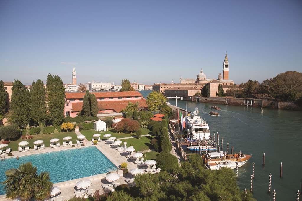 An Olympic-sized swimming pool surrounded by white sun loungers and manicured gardens, with the Venetian lagoon in the background.