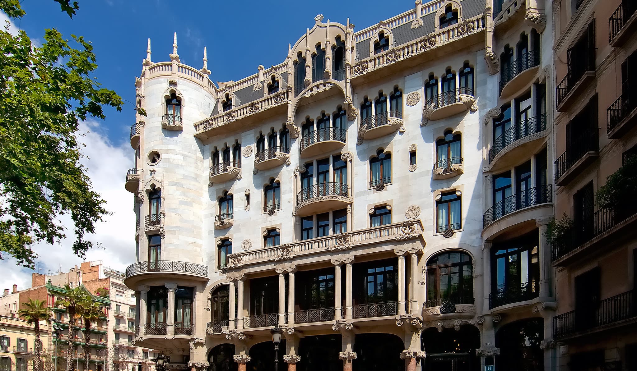 Low-angle shot of Hotel Casa Fuster's white marble facade and corner tower against a blue Barcelona sky, showcasing Domènech i Montaner's Modernista architecture.