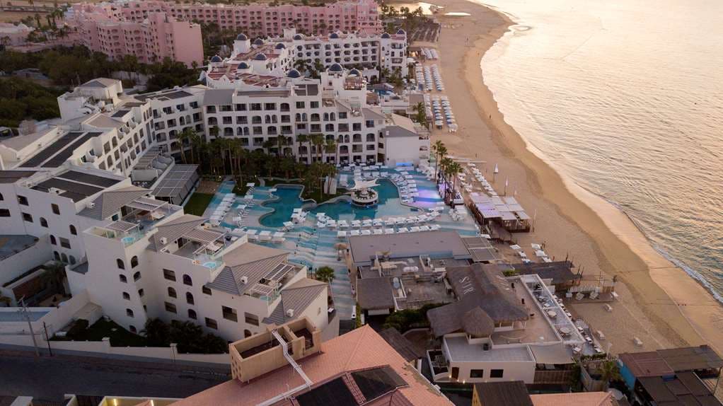 View of the ME Cabo infinity pool and Taboo Beach Club with the iconic El Arco rock formation in the background at sunset.