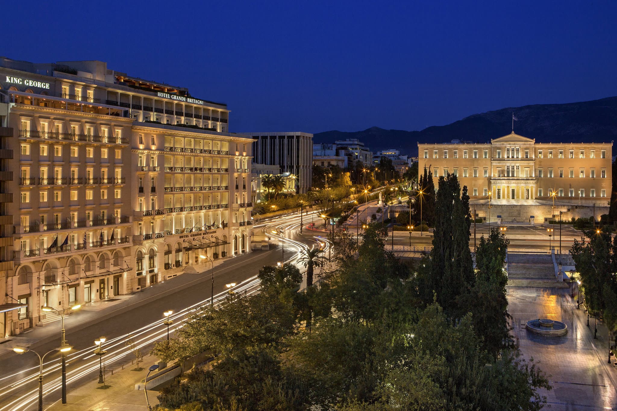 Tudor Hall restaurant terrace at night featuring elegant white tablecloth dining with a glowing view of the Parthenon and Acropolis in the background.