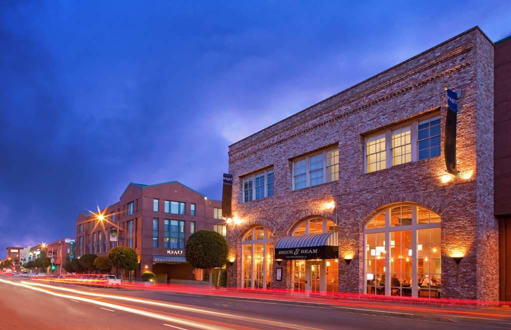 A moody, dimly lit hotel lounge featuring exposed brick walls, heavy timber beams, and a roaring fireplace, embodying the industrial-chic aesthetic.