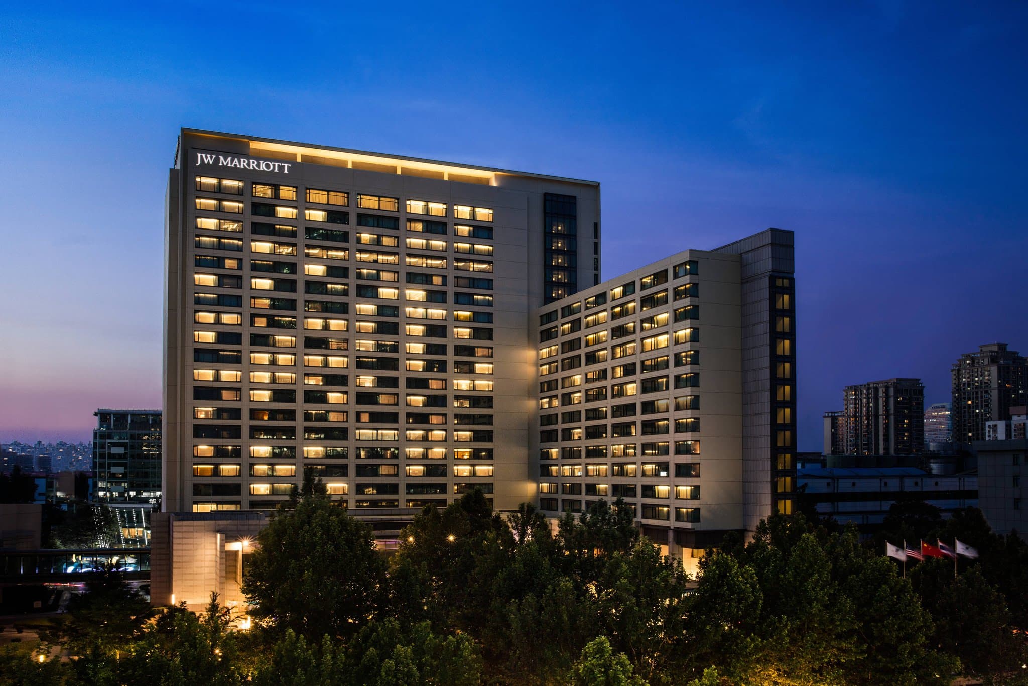 A low-angle shot of the JW Marriott Beijing exterior towering over the JW Garden, with the China Central Place skyscrapers in the background.