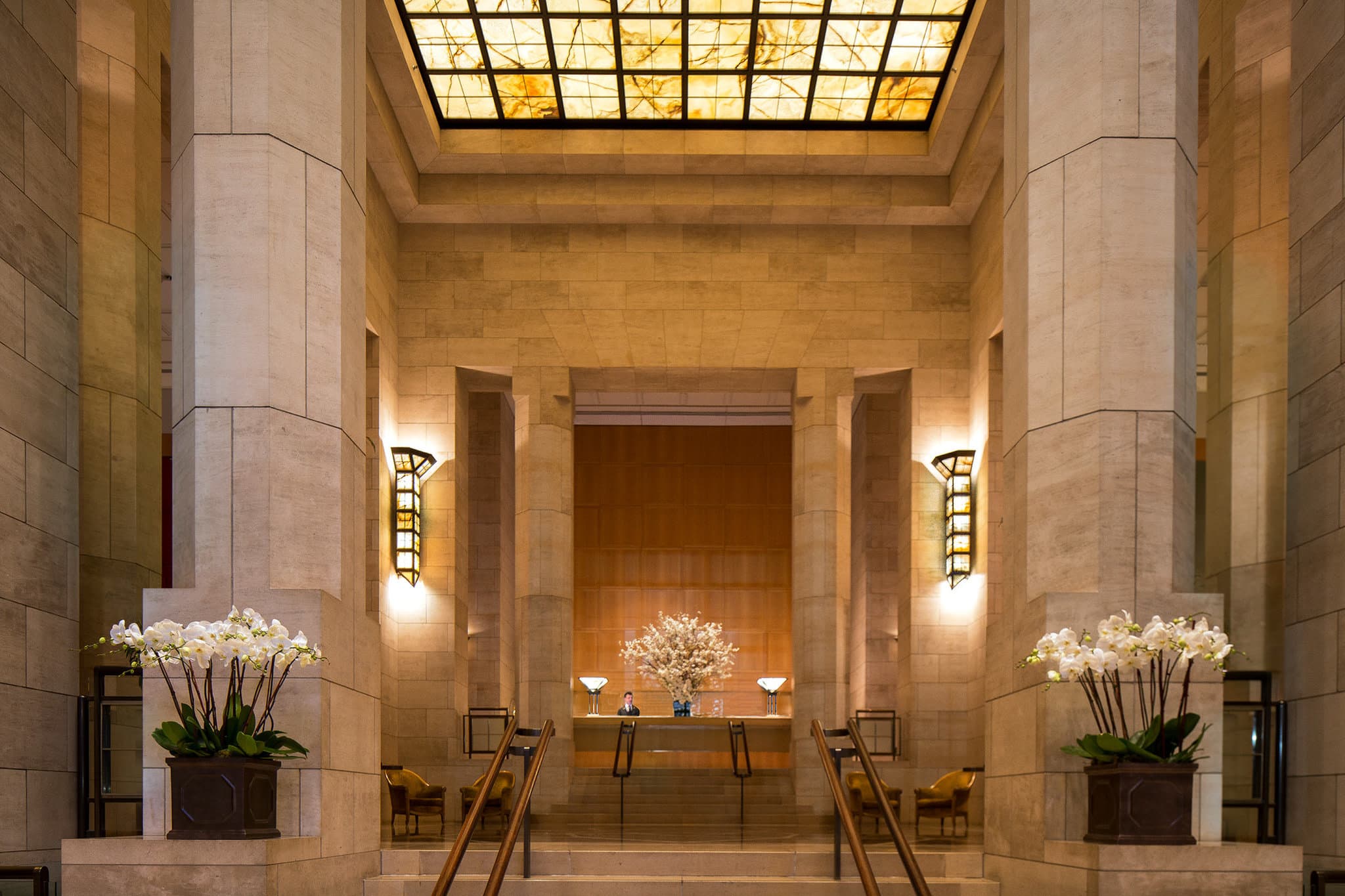 Low-angle view of the Four Seasons New York Grand Lobby featuring towering limestone pillars and a glowing onyx ceiling.