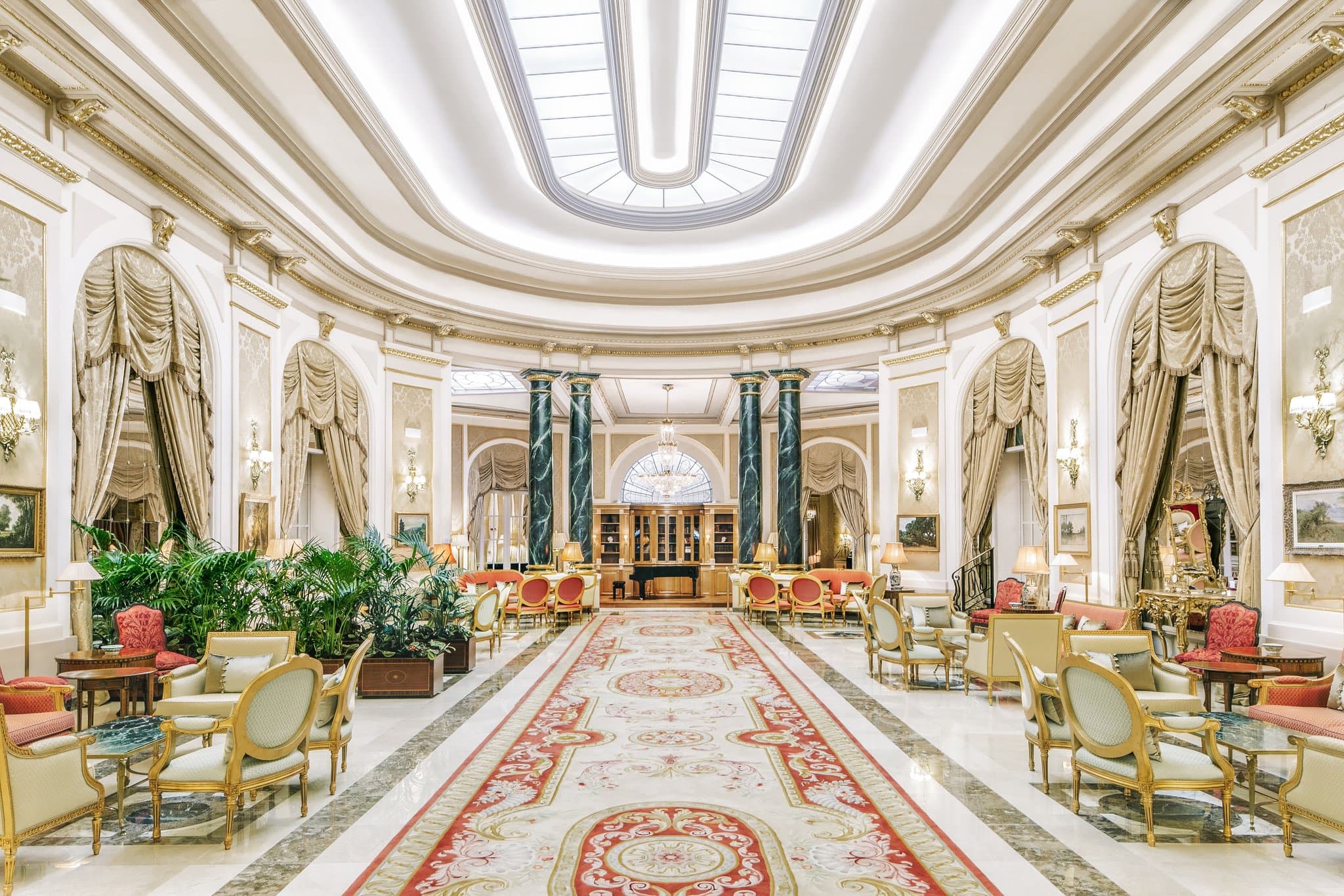 The opulent Great Hall of El Palace Barcelona featuring immense crystal chandeliers, marble columns, and velvet furniture under a stained-glass ceiling.