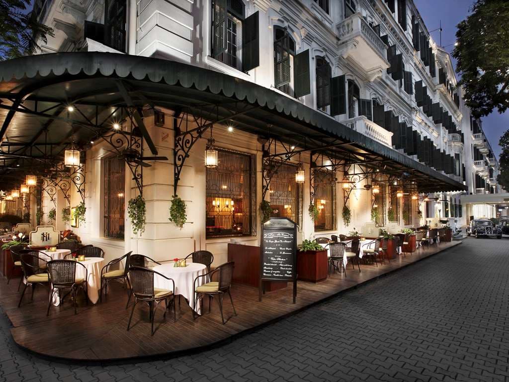 Classic white Citroën parked in front of the colonial white façade of the Sofitel Legend Metropole Hanoi, flanked by green shutters and palm trees.