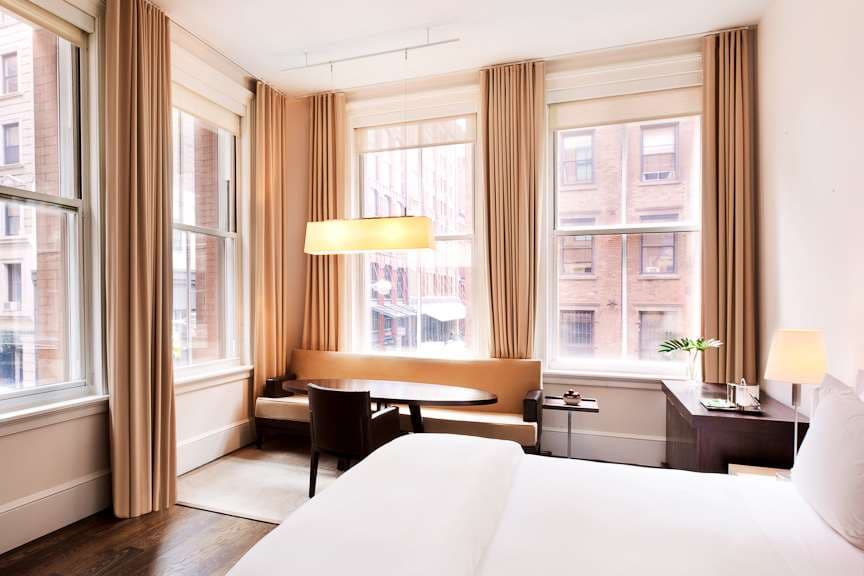 A moody, high-ceilinged loft suite at The Mercer Hotel featuring exposed brick walls, a low-profile modernist bed, and a large arched window overlooking SoHo cobblestones.
