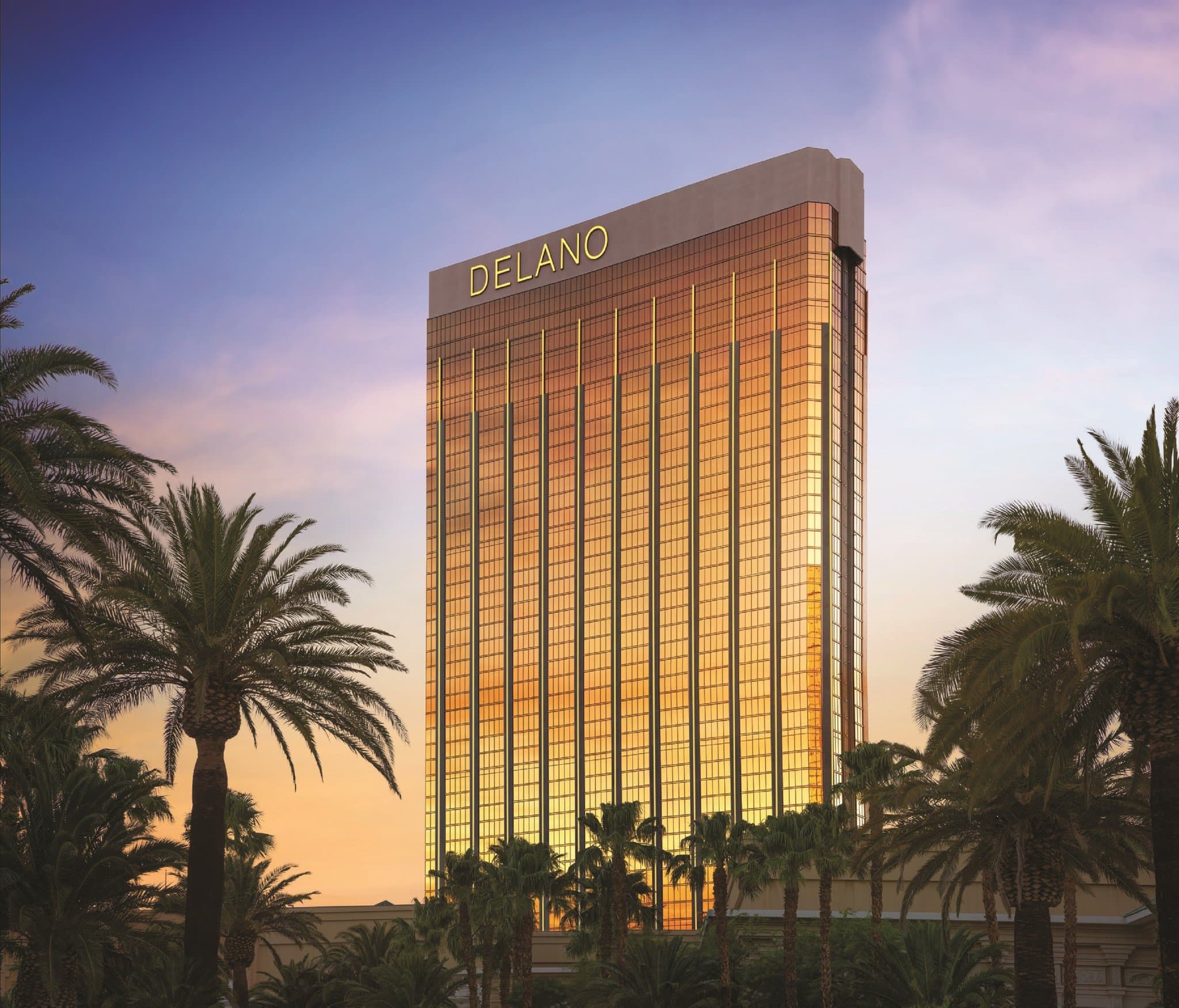 A sleek, white-walled hotel suite living room with floor-to-ceiling windows overlooking the Las Vegas Strip, featuring modern furniture and gold accents.