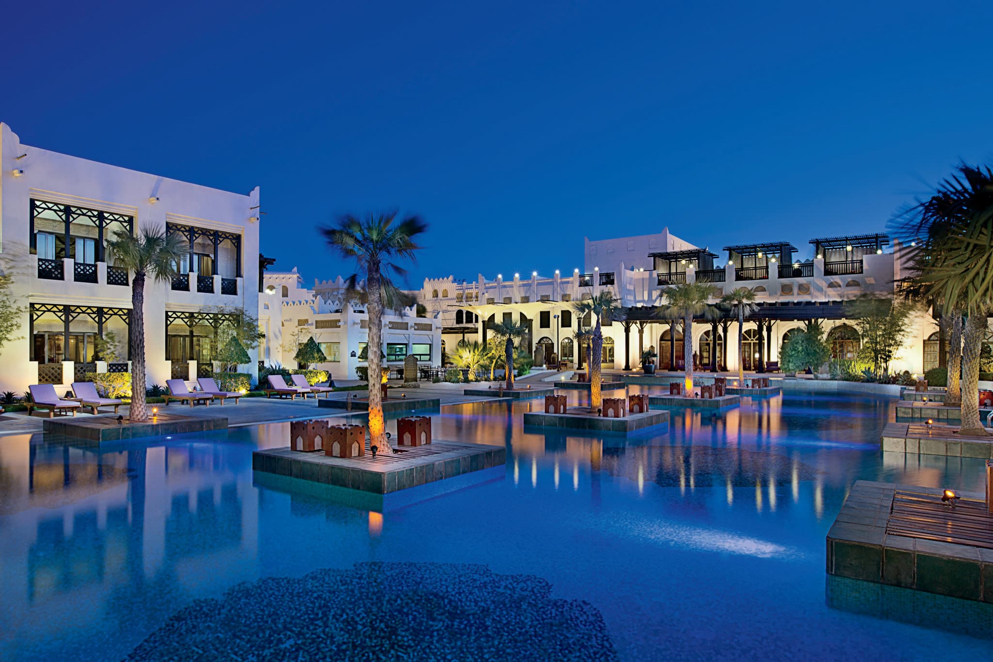 The sprawling infinity pool at Sharq Village & Spa, bordered by palm trees and traditional Qatari low-rise architecture, looking out towards the Doha skyline.