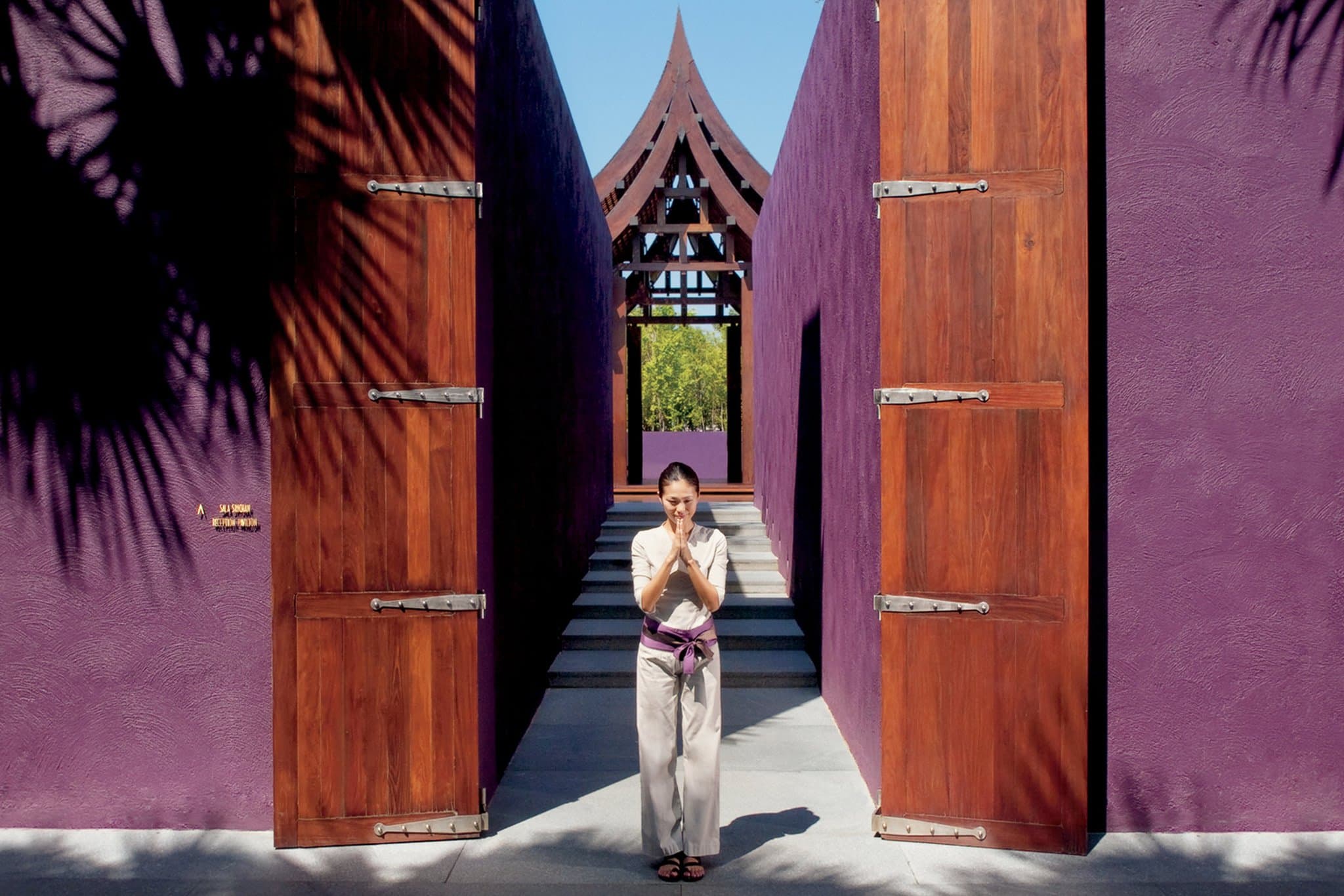 A woman in a white dress walking along a stepping-stone path surrounded by water, leading towards a traditional Thai pavilion with purple walls at Phulay Bay.