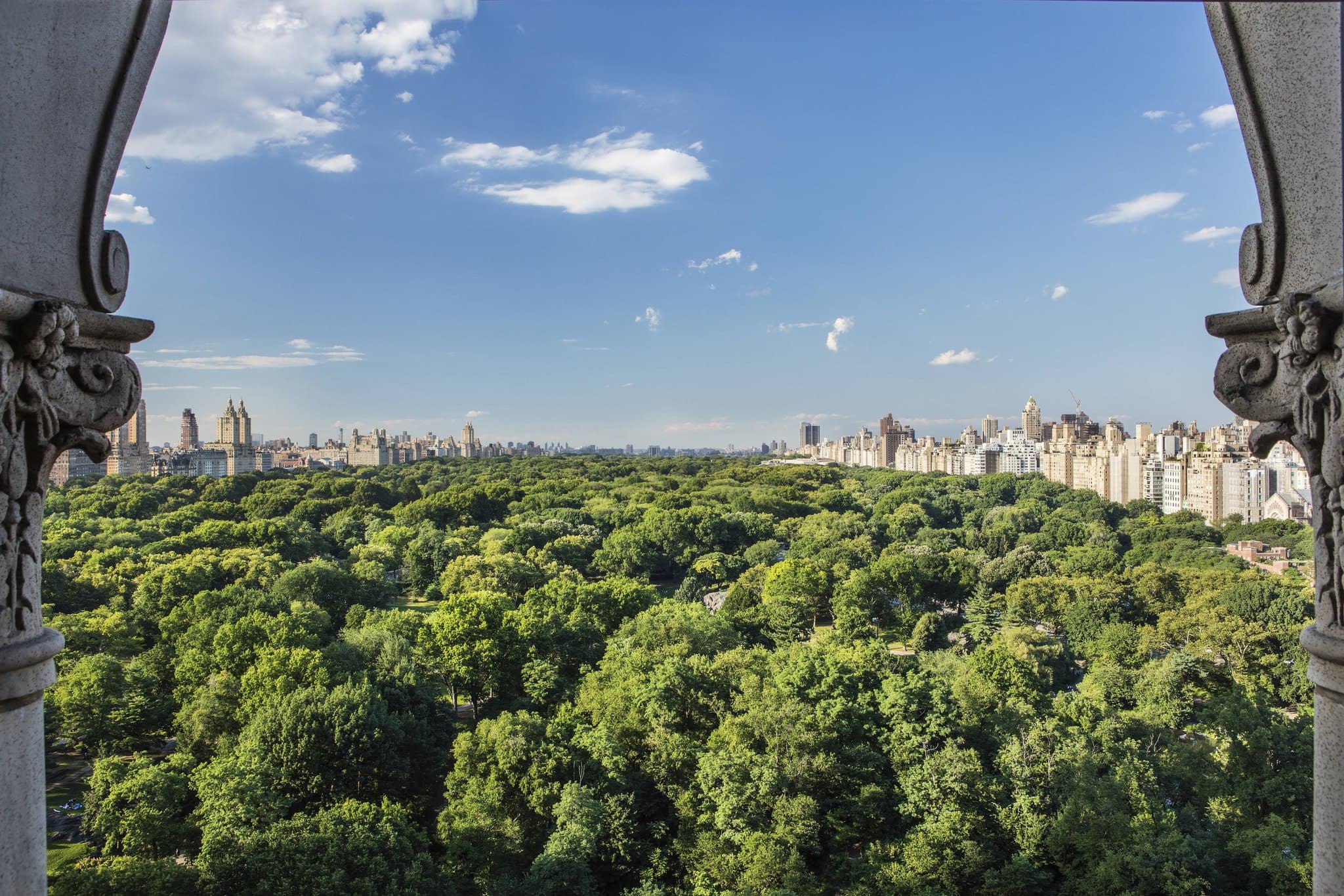 A telescope positioned by a large window in a luxury hotel suite, framing a view of Central Park's lush green treeline.