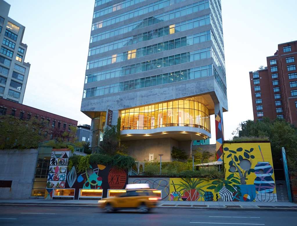 A sleek, modern hotel room with floor-to-ceiling windows overlooking the Manhattan skyline at twilight, featuring a king bed with Frette linens.
